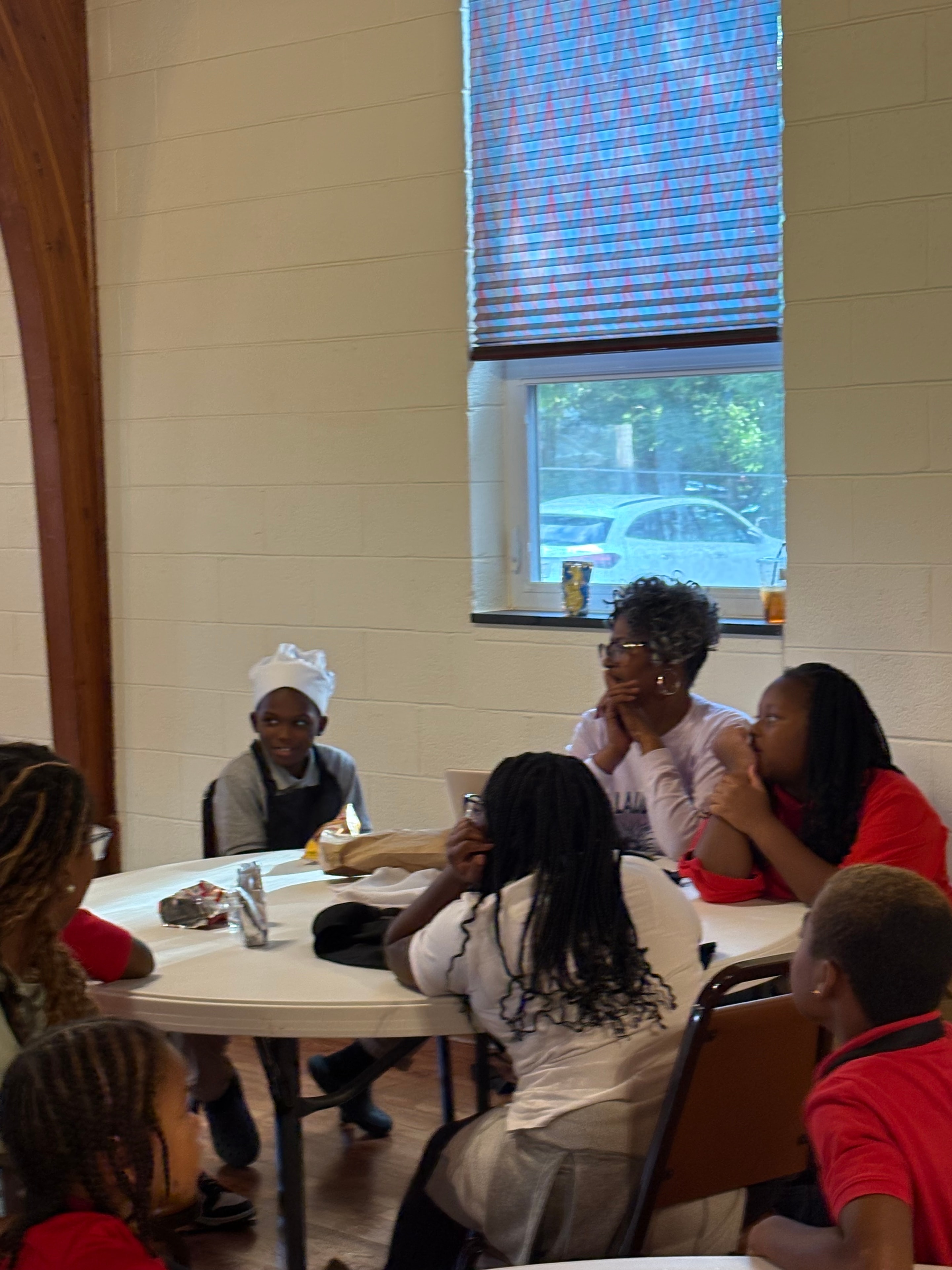 Children and adults gathered around a table while a young chef wearing a chef hat takes part in Kids R Cookin'
